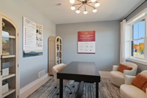 Modern office with black desk, three beige chairs, shelves, and large window letting in natural light.