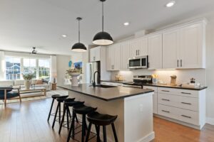 Modern kitchen with white cabinets, black island, barstools, and pendant lights, opening to a bright living area.