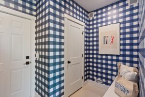 Room with blue and white checkered wallpaper, white doors, artwork, and a bench with a Vermont bag and pillows.