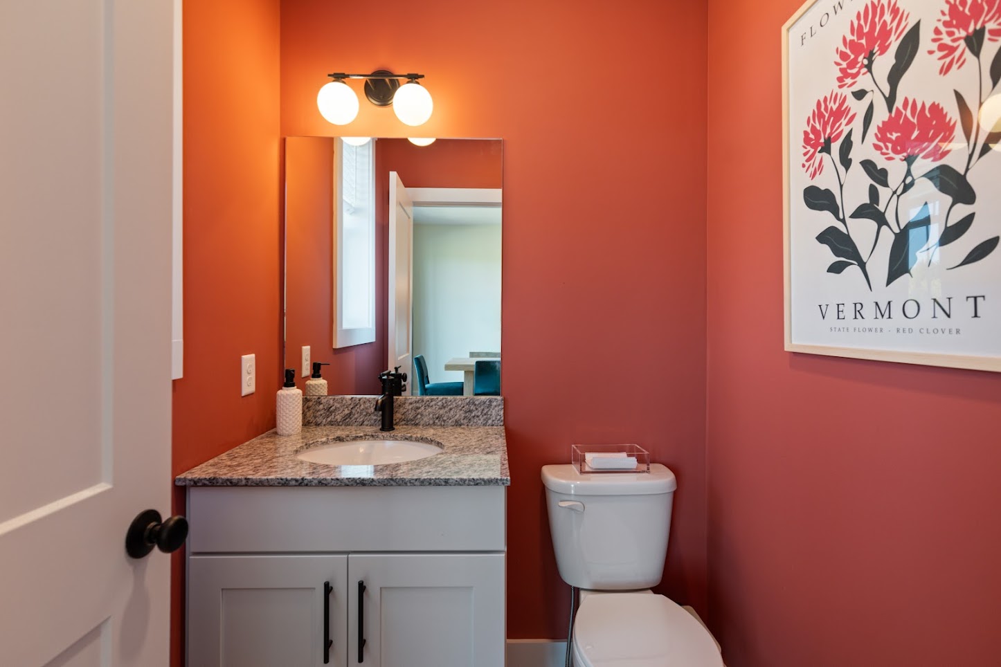 Small bathroom with red walls, granite countertop, white vanity, and a Vermont flower poster above the toilet.