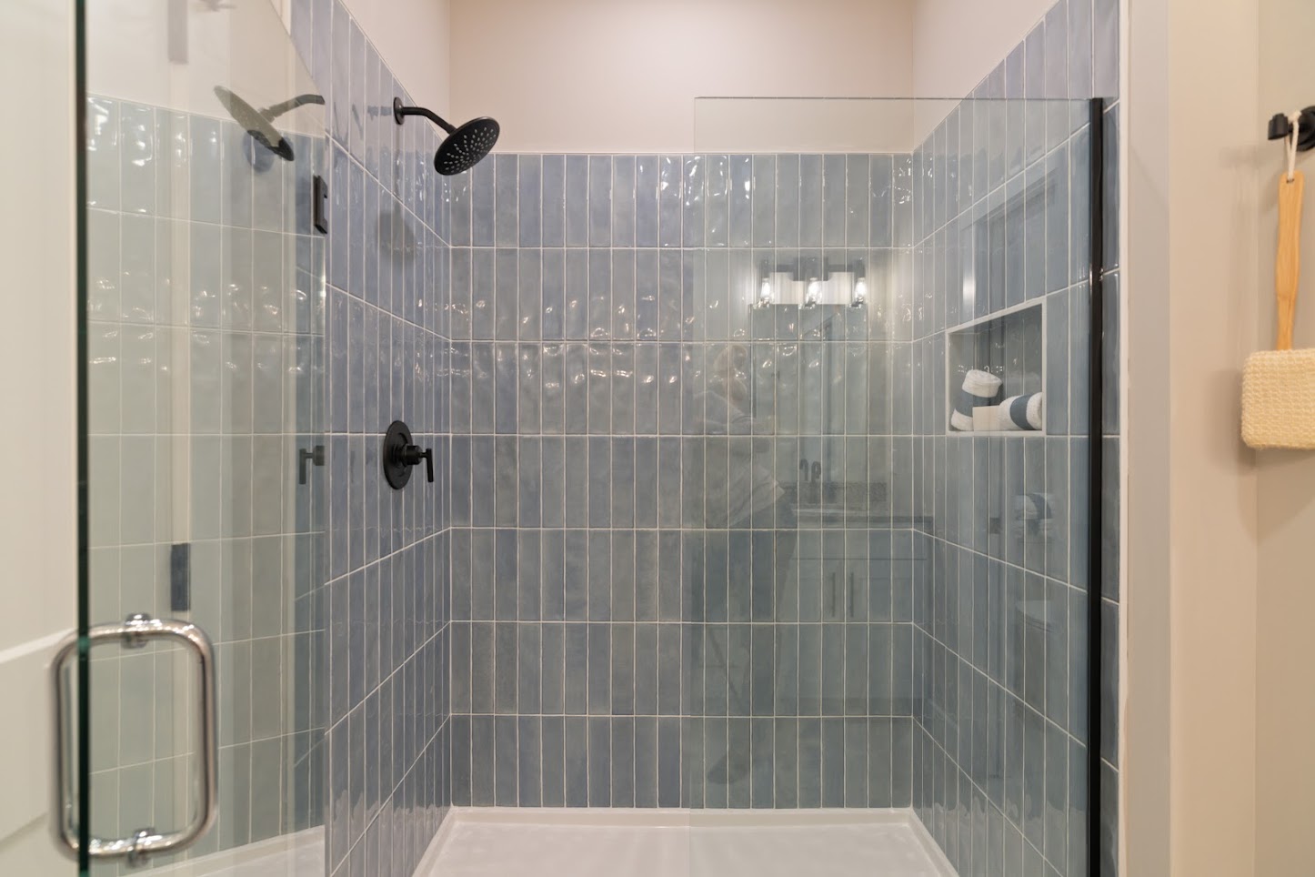 Modern glass shower with light blue vertical tiles, black fixtures, and built-in shelf holding towels and bottles.