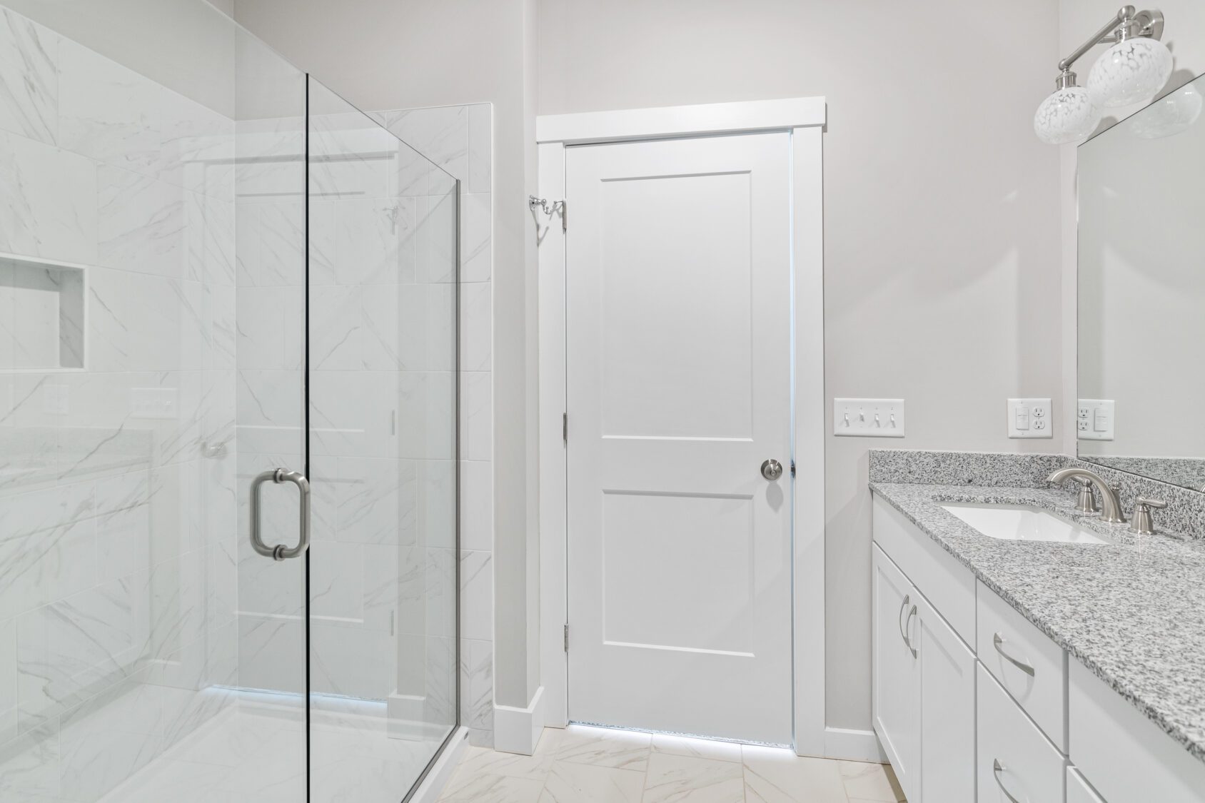 Modern bathroom with a glass shower, white door, double sink vanity, and granite countertop.