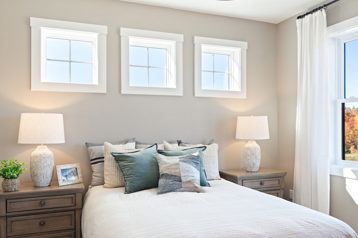 Bright bedroom with a neatly made bed, two lamps, and three small windows above the headboard.