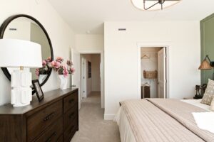 Bright bedroom with a dark dresser, round mirror, pink flowers, and an open closet with baskets and hangers.