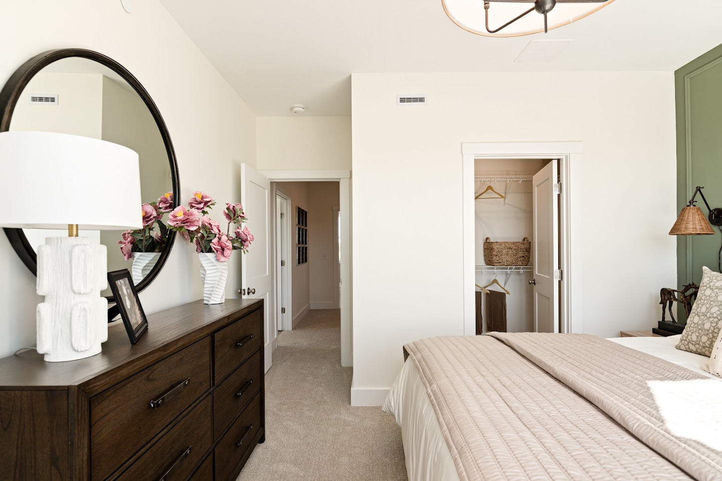 Bright bedroom with a dark dresser, round mirror, pink flowers, and an open closet with baskets and hangers.