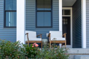 Two cushioned wooden chairs on a porch at 68 Eden Lane, nestled behind flowering bushes and a blue-sided house.