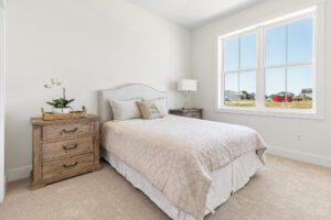 Bright bedroom at 385 Beaudry Lane with a neatly made bed, wooden nightstands, lamp, and window overlooking houses.