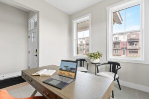 A bright home office at 68 Eden Lane features a desk, laptop, open book, two chairs, and large windows with neighborhood views.