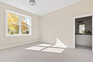 Bright, empty beige bedroom at 68 Eden Lane with large window, carpeted floor, and view into a bathroom with vanity.