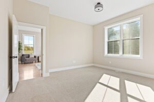 Bright, empty room at 68 Eden Lane with beige walls, carpet, large window, and open door to a gray chair in the sitting area.