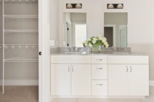 White bathroom vanity with two sinks, large mirrors, and a vase of flowers in the 68 Eden Lane open closet with shelves.