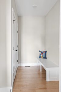 A minimalist hallway at 68 Eden Lane features a white bench, wooden floor, and colorful pillow.
