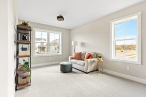 Bright living room at 68 Eden Lane with a beige sofa, ottoman, side table, shelves, and sunlit large windows.