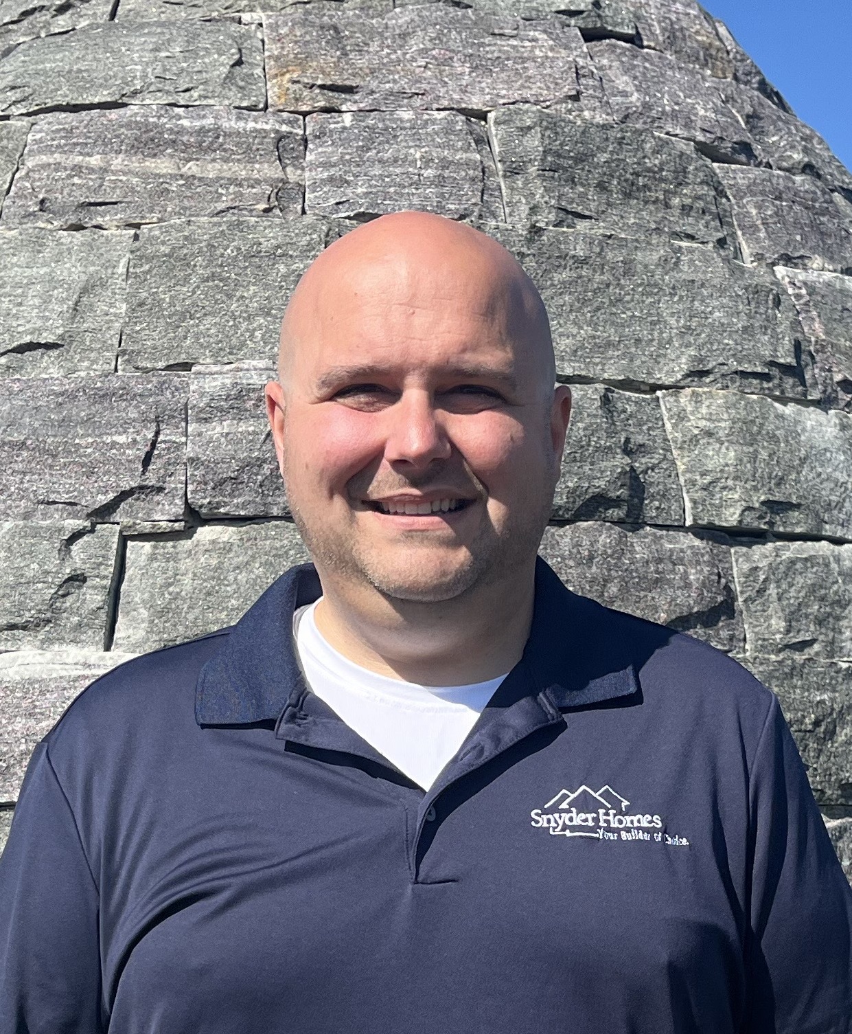 A smiling man in a navy Snyder Homes shirt stands in front of a stone wall on a sunny day.